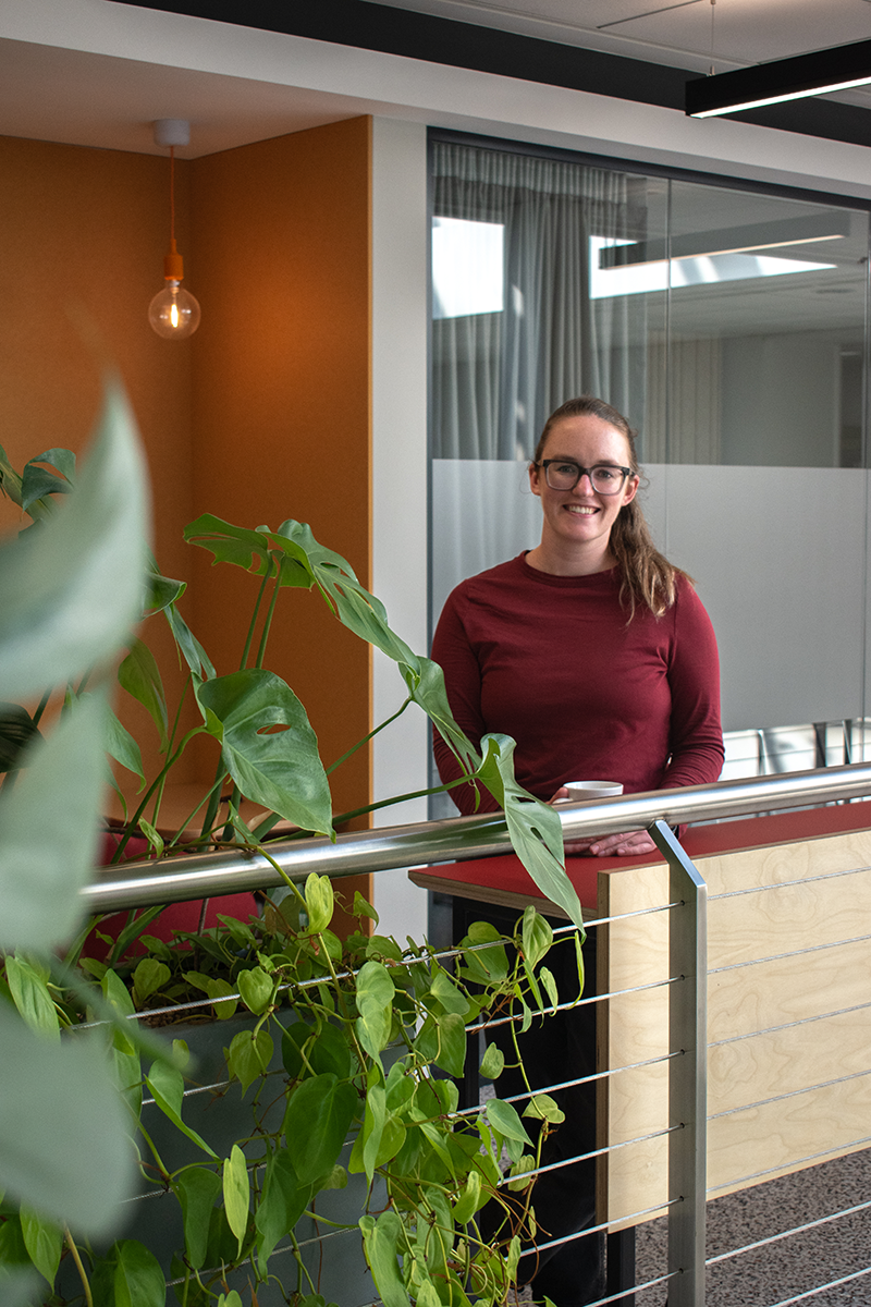 Catherine Sinclair, Head of Research, smiling while wearing a red shirt, with plants surrounding, zoomed out portrait image