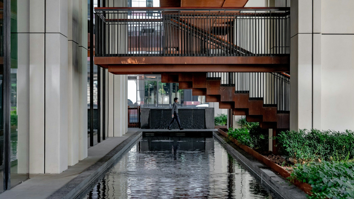 A person walking beneath some copper steps, with a body of water in front, surrounded by the Brutalist architecture of the Barbican