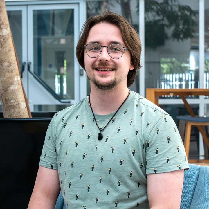A young adult with medium-length hair and glasses smiles at the camera while seated indoors. They are wearing a light green patterned T-shirt and a necklace, with modern furniture and large windows in the background.