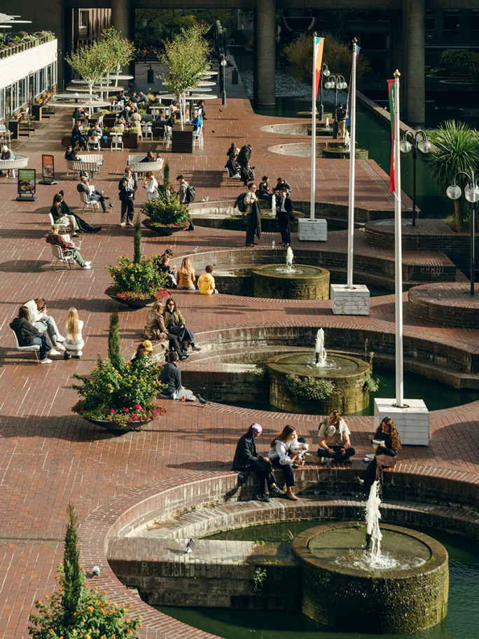 A view of the Barbican social area from a balcony. There are four circular fountain features and multiple people sitting in groups talking.