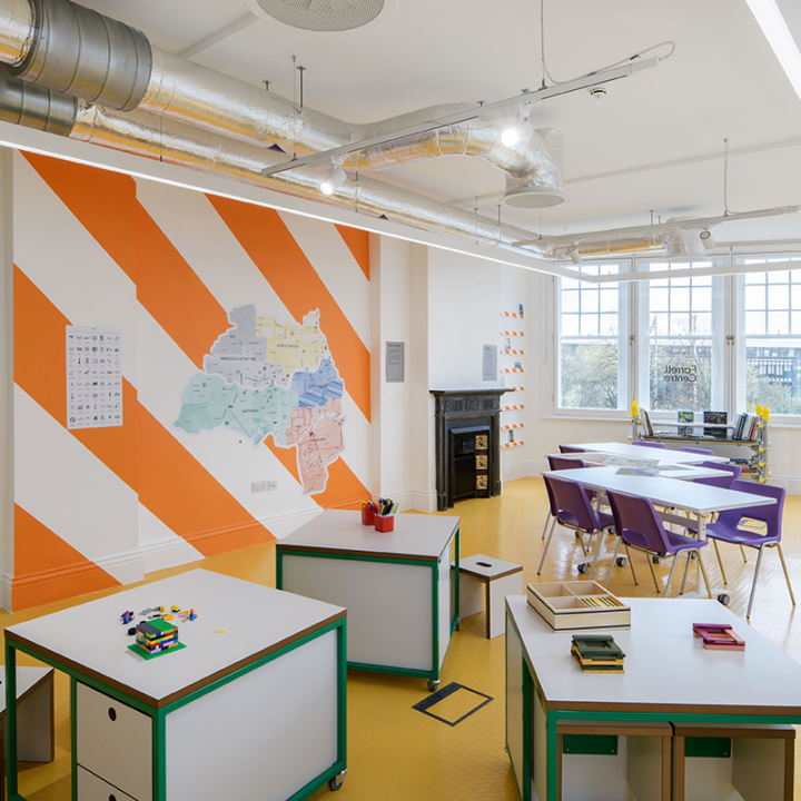 Inside the Farrell Centre. There are multiple workspaces with green edges and in the far right, two longer tables with purple chairs. The wall on the left has orange and white diagonal stripes and a map of Newcastle.