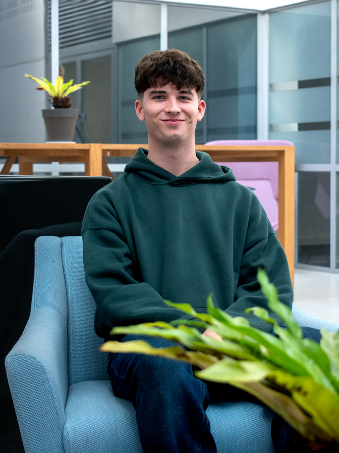 A young adult with short brown hair and a green hoodie jumper sits on a blue sofa seat and smiles at the camera