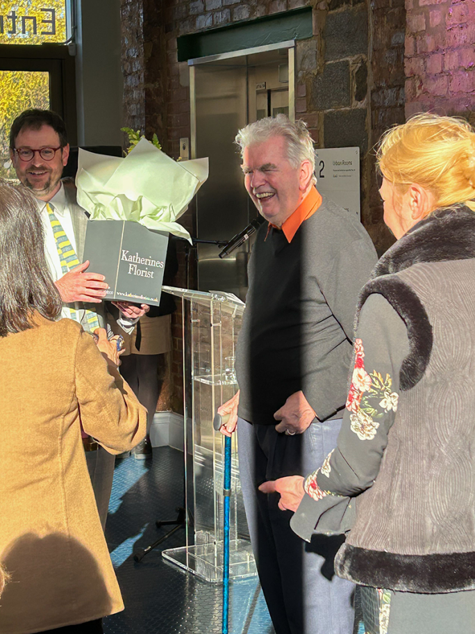 An Image of Sir Terry Farrell smiling while wearing blue trousers and a black jumper with orange collar. He is surrounded by two women and a man with glasses who is handing him some flowers in a bag labelled 'Katherine's Florist'