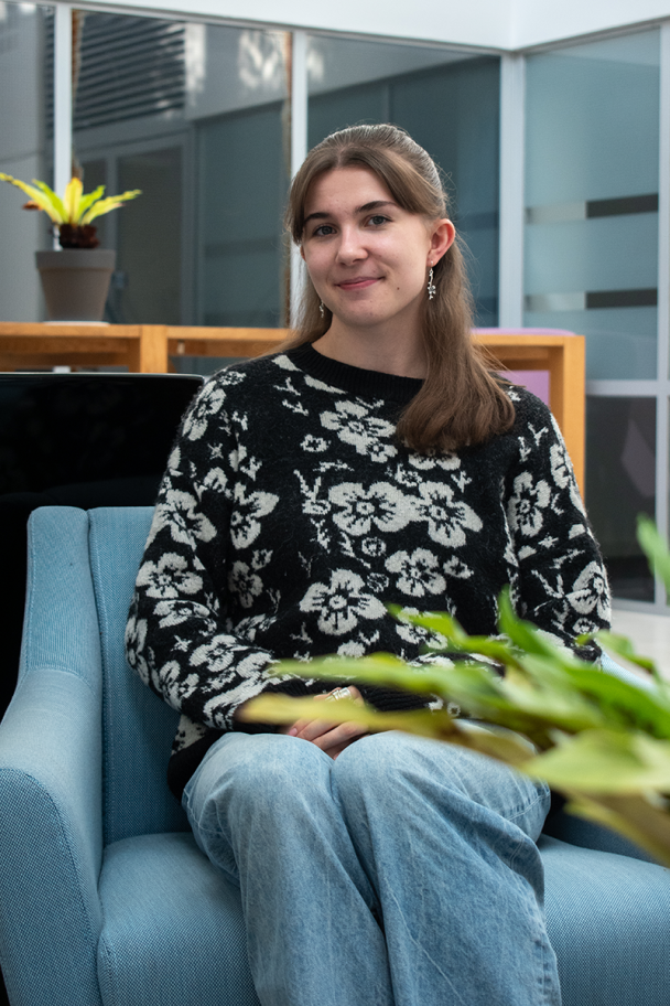 A young woman with medium length brown hair and a parted fringe sits on a blue sofa smiling at the camera, she wears a black jumper with white patterned flowers and blue jeans