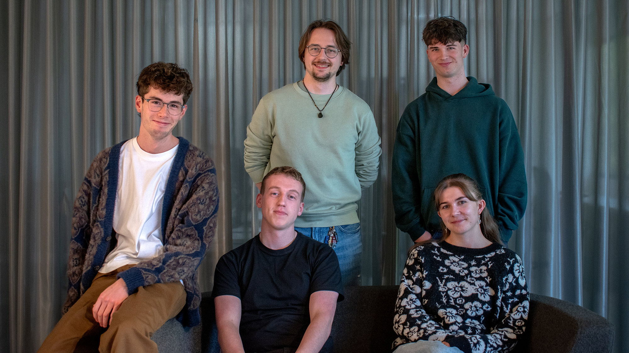 A group of five young adults sit and stand together in a casual indoor setting, smiling at the camera. Two people are seated at the front while three stand behind them, all dressed in relaxed, comfortable clothing.