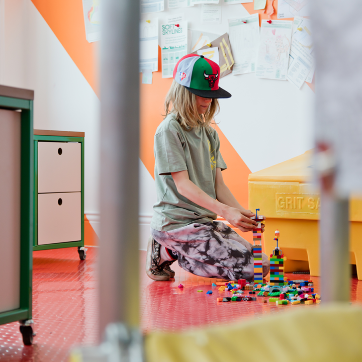 A young boy wearing a faded green t-shirt and tie-dye patterned trousers kneels on the floor building a lego tower. He is wearing a white, green and red cap with a red bull on it.