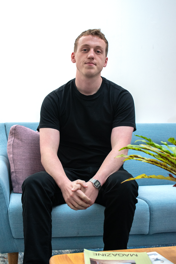 A young professional sits on a blue sofa with a pink cushion, he wears a black t-shirt and black jeans and is holding his hands together between his knees