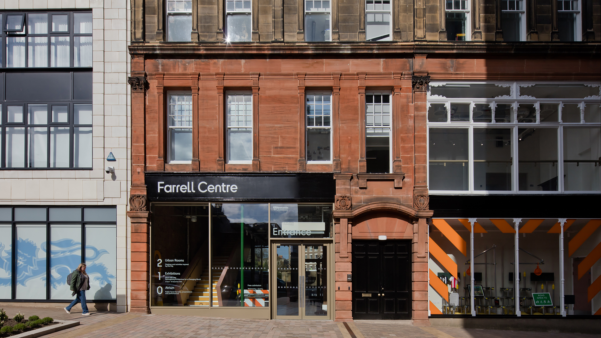 A street view of the Farrell Centre in Newcastle Upon Tyne, a Victorian looking tree-story terrace building with grey and red brick. There is a woman walking past on the street below.