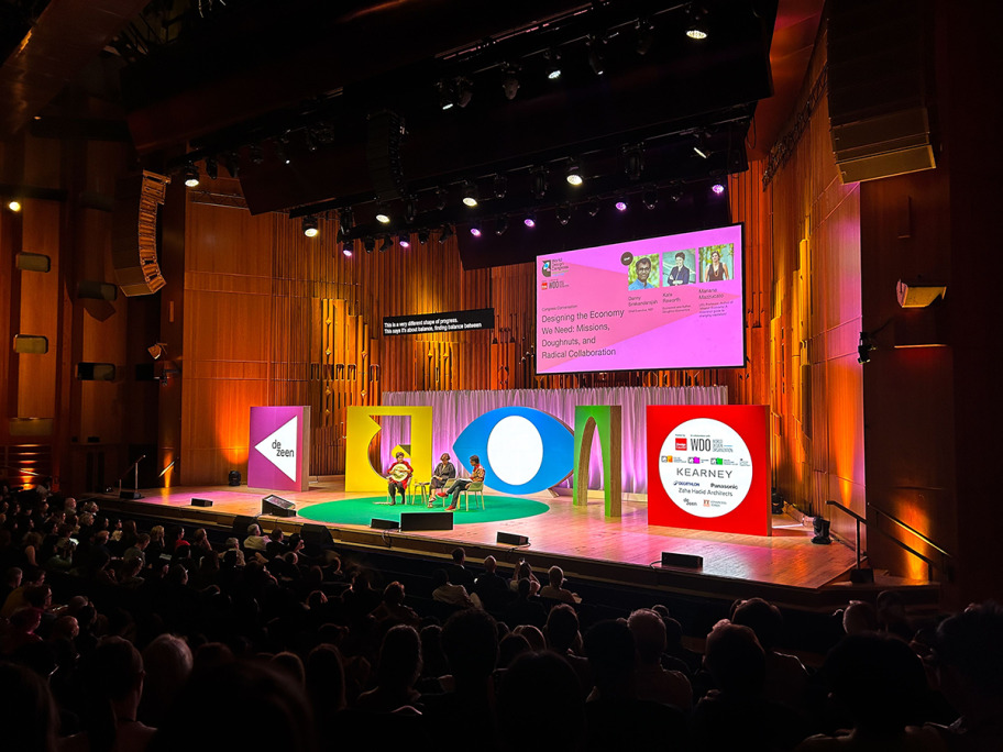 Kate Raworth and Mariana Mazzucato are panel speakers. They sit on a stage with the presenter, and behind them are bamboo structures on the walls and large colourful shapes.