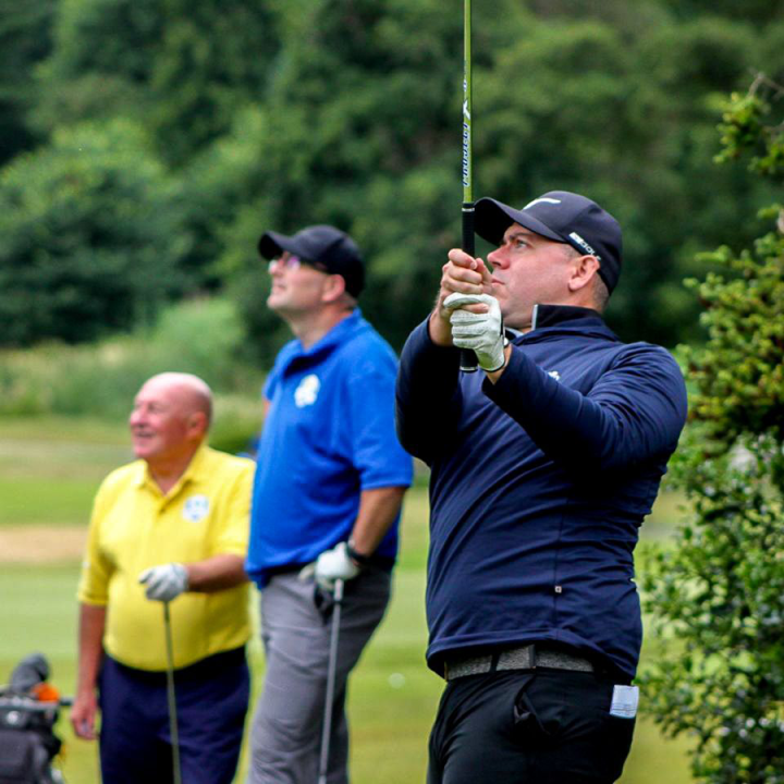 Three men in varying shades of blue shirts, one man in a yellow shirt, playing golf.