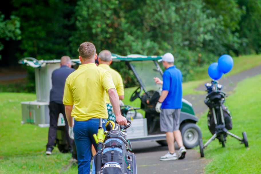 Four men walk towards a golf buggy. The two closest to the buggy wear black and blue shirts and the two further away wear yellow shirts. One man is dragging a bag of golf clubs behind him.