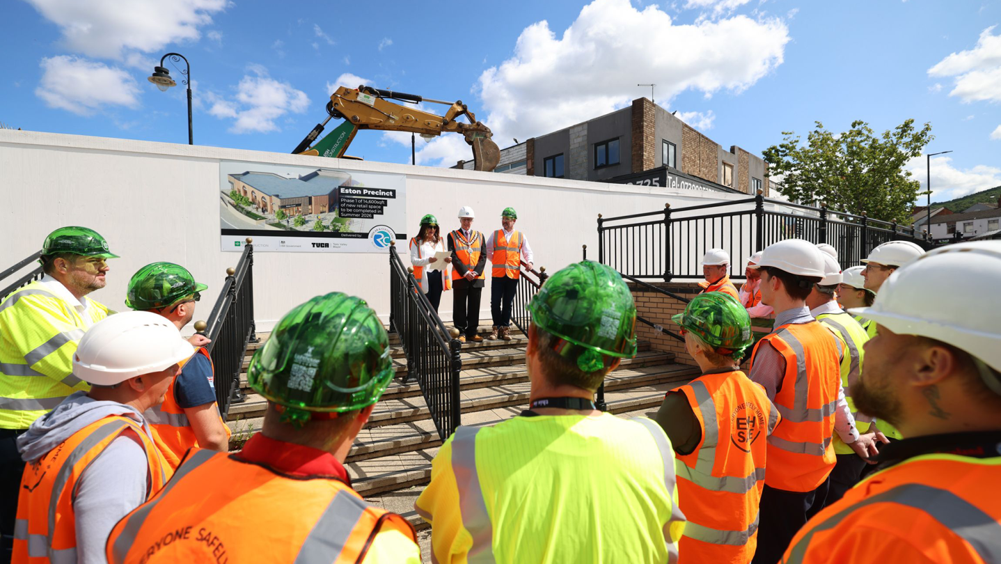 One woman and two men stand at the top of some steps addressing an audience of contractors, consultants and architects. They are all wearing high-vis jackets and hard hats.