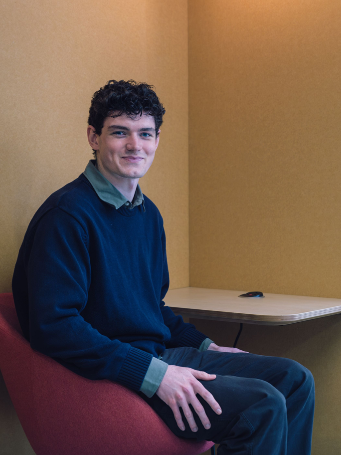 Euan Ellis is sitting on a red chair smiling at the camera, he wears a blue jumper with a green collar and grey trousers