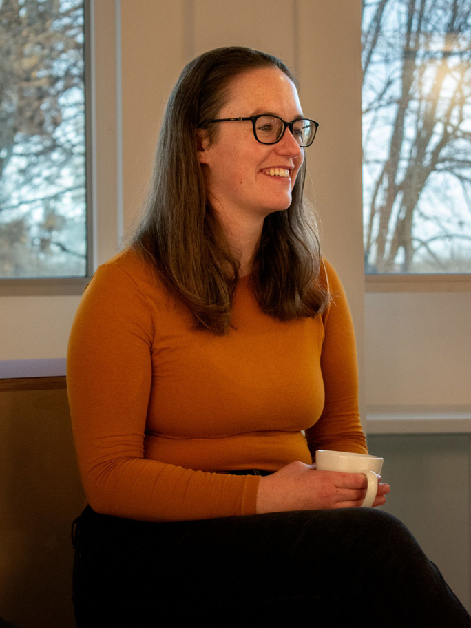 Catherine Sinclair smiles into the distance, she wears a yellow long-sleeved top and has a coffee mug in her hand
