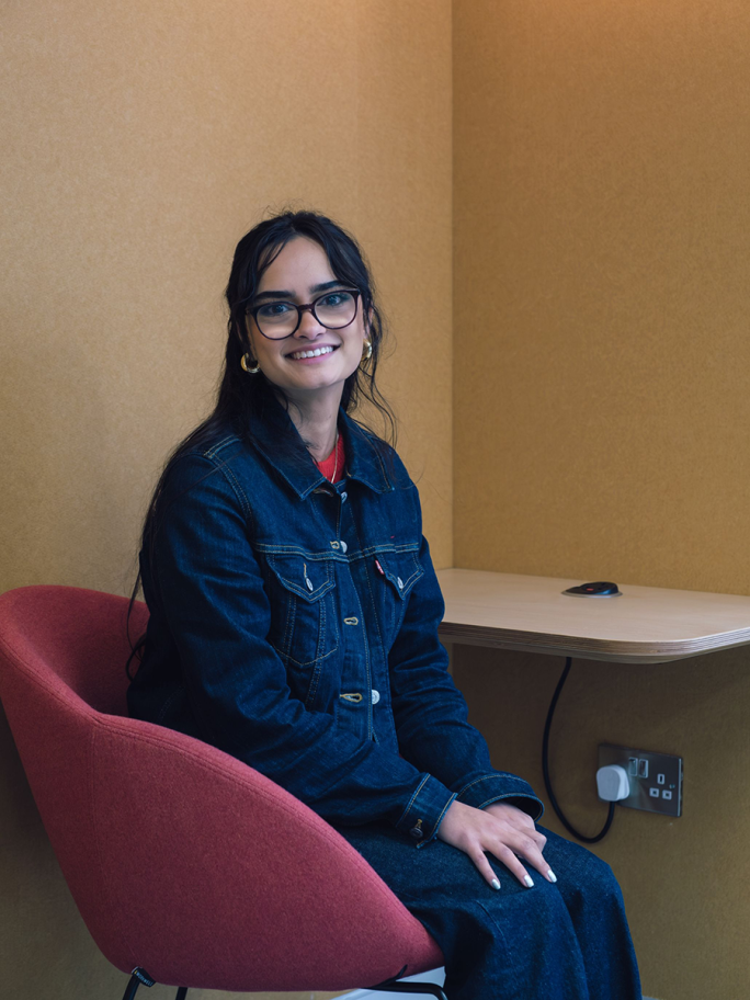Hannah Rolfe sits on a red chair facing the camera, she is wearing glasses and a denim jacket & jeans