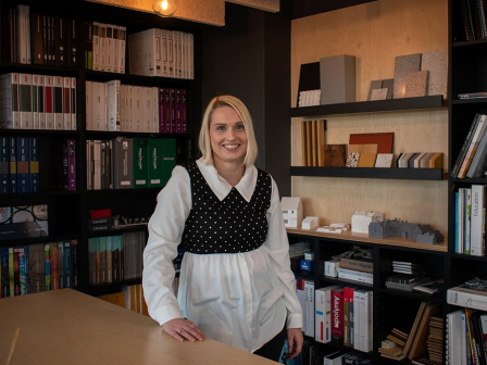 Image of Donna Khan posing and smiling in front of a Library nook. She wears a white shirt with a black and white cropped polka dot jumper over the top.