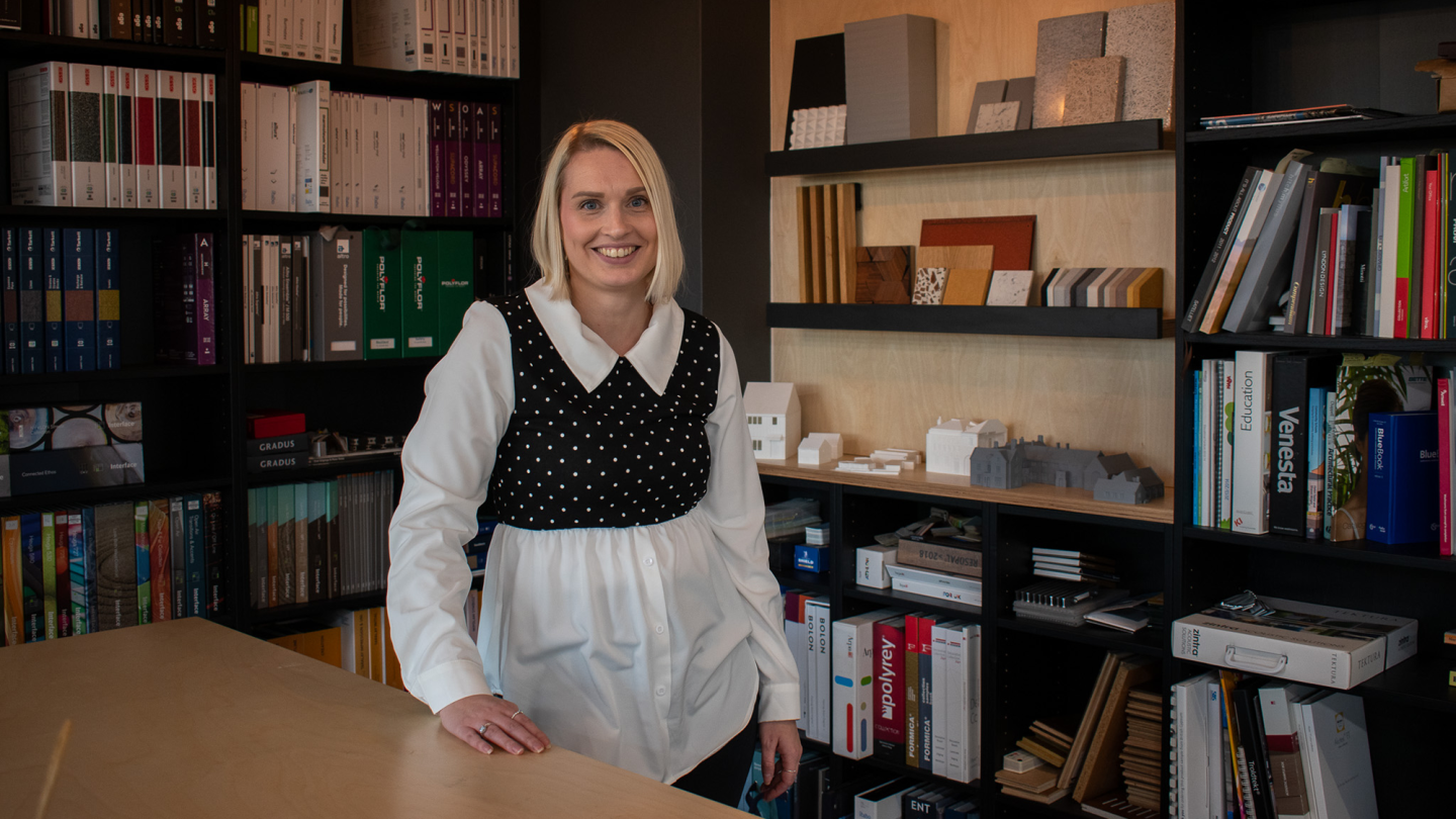 Image of Donna Khan posing and smiling in front of a Library nook. She wears a white shirt with a black and white cropped polka dot jumper over the top.