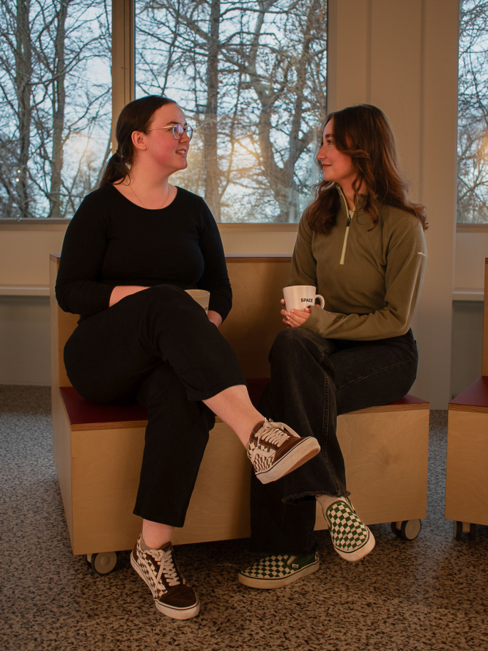Jade Atkins, wearing a long sleeved black top and black jeans smiles at Brook Wilkinson, who wears brown trousers and a sage green sports jumper