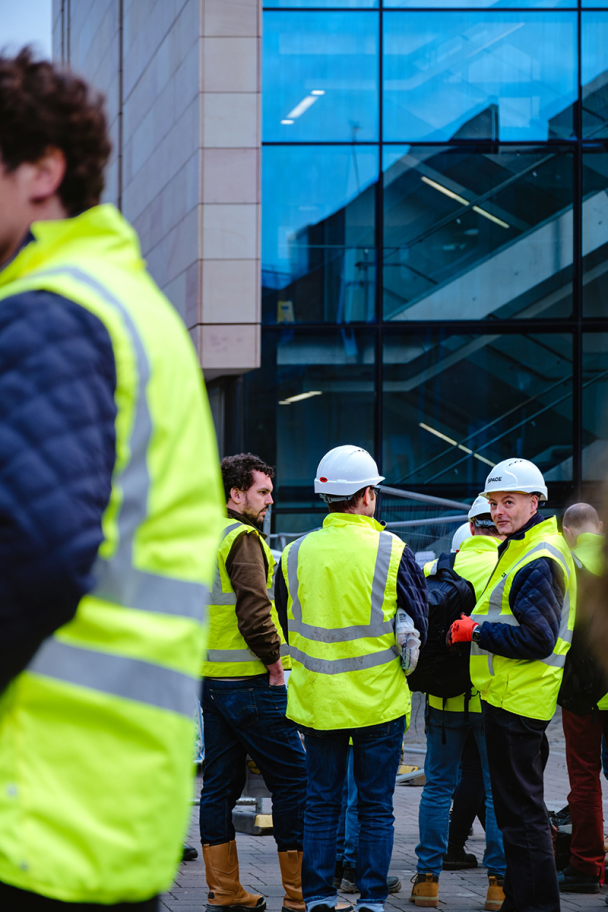 A zoomed out image of 3 men in high vis jackets and hard hats, one is looking directly into the camera as the others look away