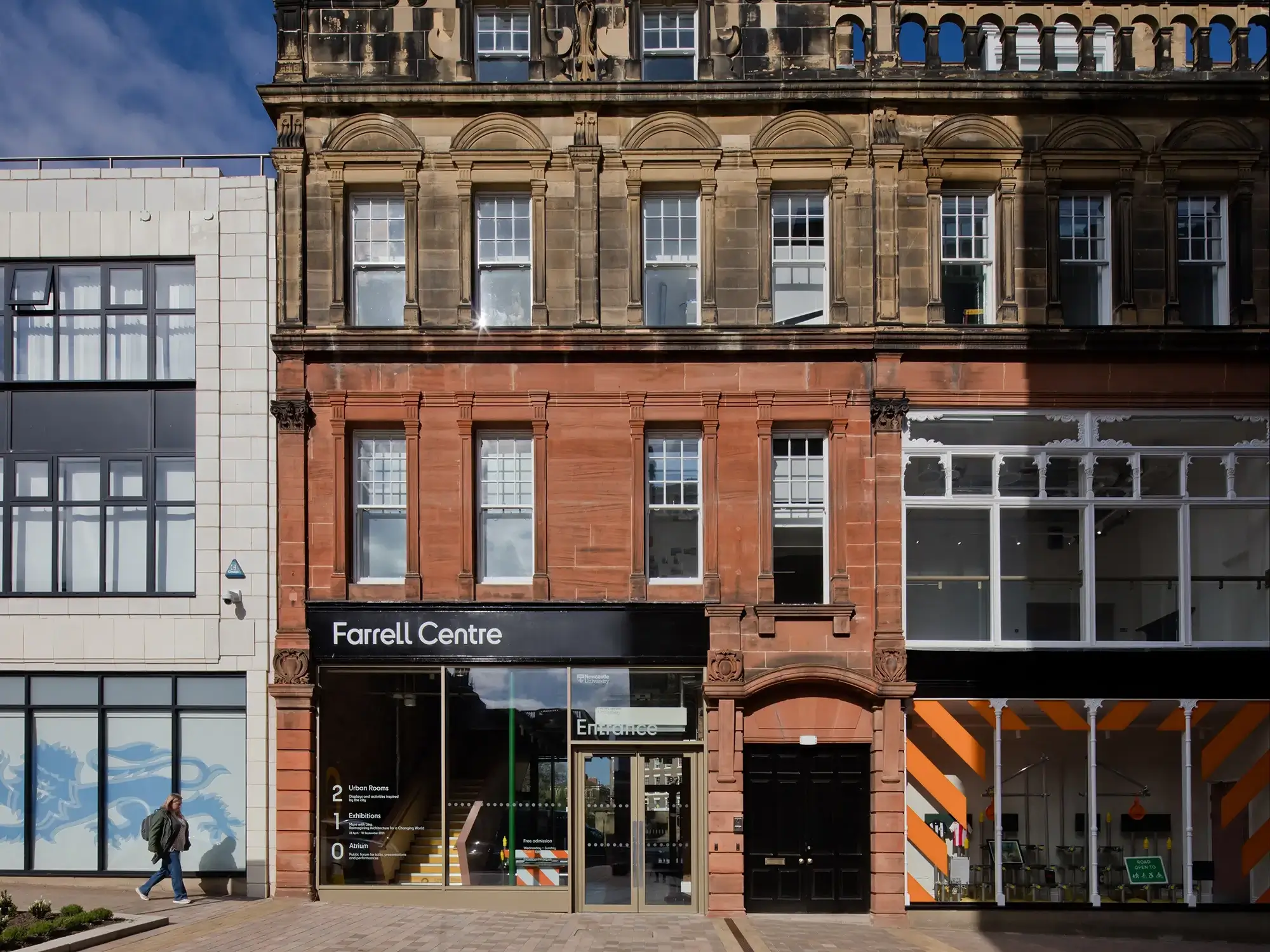 A street view of the Farrell Centre in Newcastle Upon Tyne, a Victorian looking tree-story terrace building with grey and red brick. There is a woman walking past on the street below.